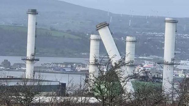 Ballylumford power station  - white chimneys, one is visibly damaged and bent, dwellings and mountains visible in background