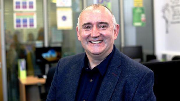 Cormac is smiling happily, wearing a dark blazer and shirt. Behind his is an office space. 