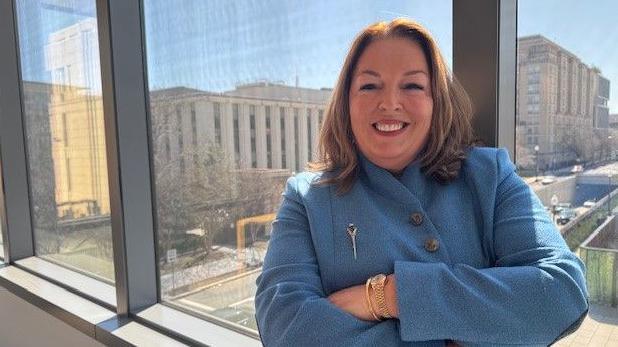 Tina McKenzie has long dark hair and is smiling at the camera. She is wearing a light blue coat with a silver broach and has her arms folded. A gold watch and bracelet are visible on her left arm. She is standing at a window in washington DC with buildings in the background.