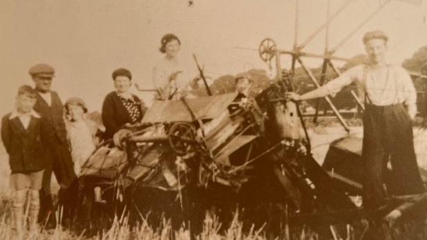 A sepia-tinged photo of a group of adults and children posing with farming equipment in a field in the 1930s. They wear typical clothes for the time.