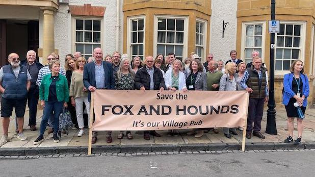 Men and women gather outside a white building holding a banner that says "Save the Fox and Hounds. It's our village pub"