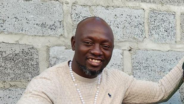 Zambian MP Emmanuel Banda wearing a white sweater and beaded necklace, in front of a grey wall