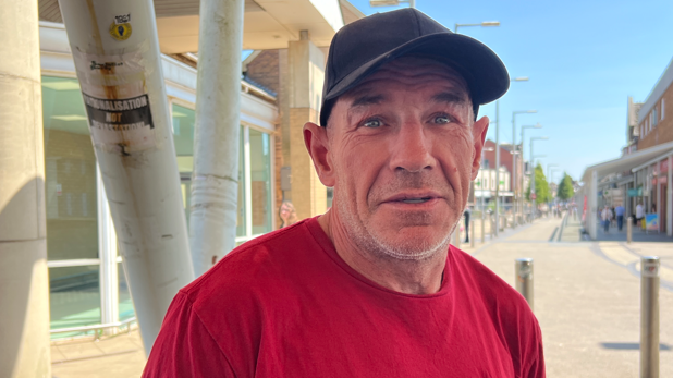 A man wearing a red t shirt and black baseball cap stood in front of a high street on a sunny day.