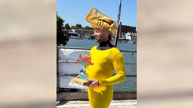 A man in skin-tight yellow clothing with a golden chef's hat on his head. He is carrying a winner's trophy