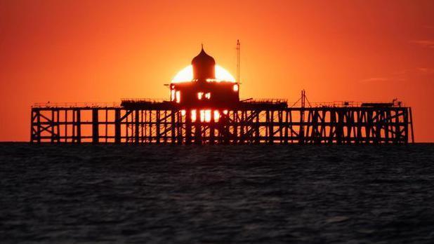 A construction in the middle of the ocean with the sun going down behind it with an orange sunset 