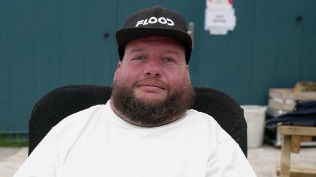Karl Flood looking directly to the camera. He is sat in a wheelchair and has a white T-shirt on and black hat.