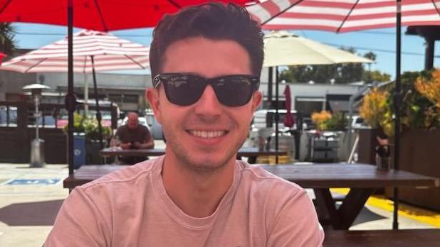 Mason Leposavic smiled while wearing sunglasses, sitting in a spot with picnic tables and umbrellas