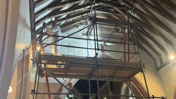 Scaffolding is in place inside the church next to the wall with the painting with extra lighting in place for it to be studied. A man in overalls stands on the higher platform. Wooden beams can be seen in the roof of the church.