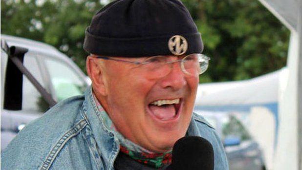 Dewi Morris laughing into a microphone, wearing a beanie hat, and glasses