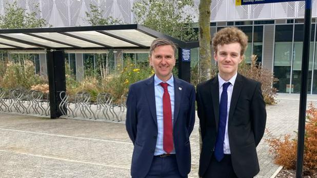 Andrew Pakes and Sam Carling face the camera with their hands behind their backs. They both wear dark coloured suits and ties. In the background is a cycle park and glass fronted building.