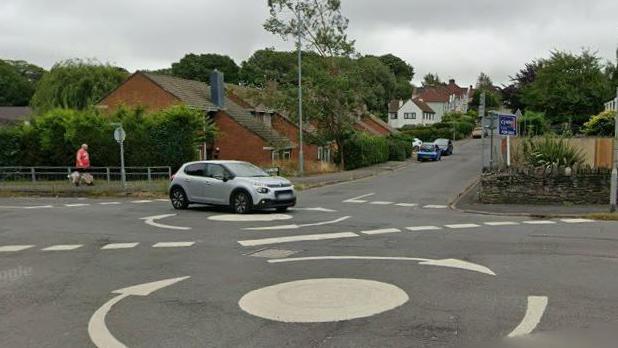 A double mini roundabout on a suburban street. Houses and parked cars can be seen. A grey car is crossing the roundabout.