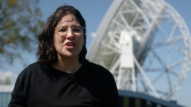 Dr George Dransfield, with shoulder length black hair, glasses and a black jumper, stands in front of the huge white dish and scaffold structure of the Lovell Telescope