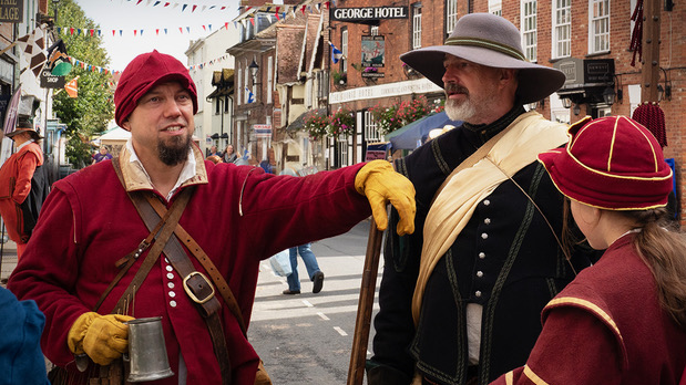 A group of people dressed in 17th-Century attire gather on a cobbled street outside the George Hotel, taking part in a historical re-enactment. They are wearing wide-brimmed hats, doublets, and gloves, with one individual holding a metal tankard. Bunting and hanging flower baskets add to the festive atmosphere in this picturesque market town setting.