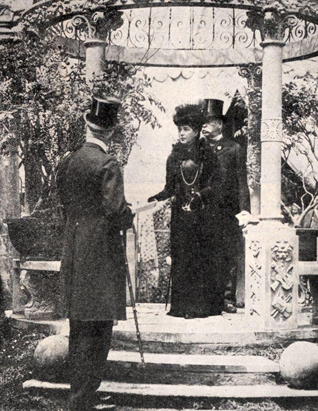Black and white photo of Queen Alexandra standing on a bandstand, and there are two other, both wearing suits and top hats.