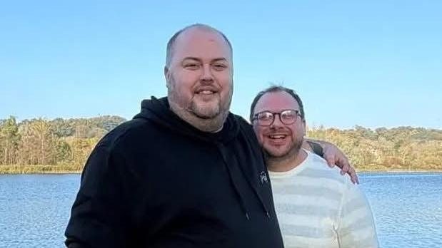 Ben Cronin stands closely with his partner as they pose and smile for a photo in front of a lake. Ben wears glasses and a white and grey striped long-sleeved top. Andrew has a shaved head and a short beard. He wears a black hoodie.