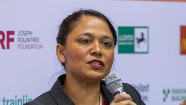 A woman with dark hair is holding a microphone, and wearing a blazer.