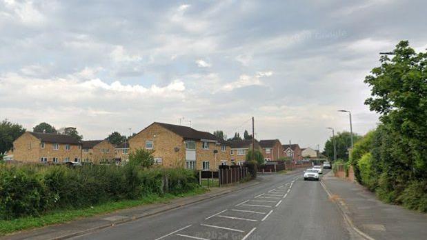 A road with three cars travelling up the carriageway on the right. A housing estate can be seen on the left, bordering a large hedgerow.