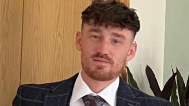 Evan Thomson is wearing a navy checked suit with a white shirt and Paisley pattern tie. He has thick brown hair and a short beard.