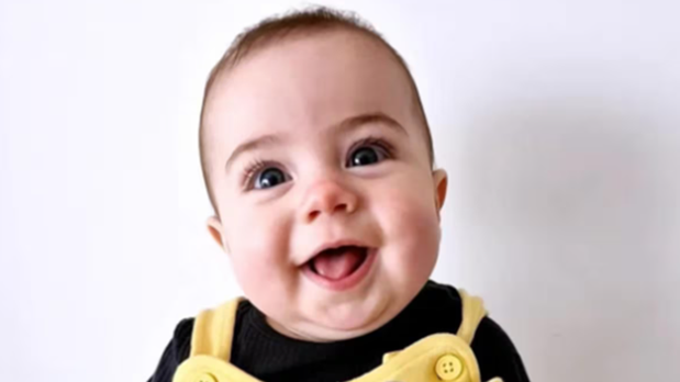 Baby girl, with big blue eyes smiling into the camera, wearing a yellow dress and black long sleeve top. 