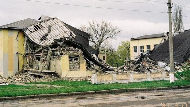 A large collapsed building next to a road. It appears to be a two-storey structure that has collapsed in the middle due to being struck.