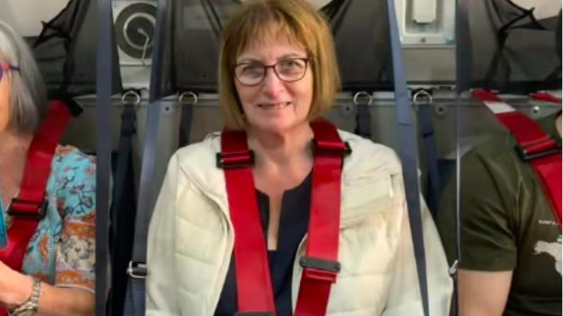 An elderly woman with a bob sat in a plane seat with two red seatbelts coming over her shoulders down across her torso. She smiles at the camera