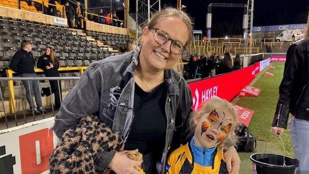 A woman aged around 40 with her arm around a young girl wearing tiger-themed face paint. They are stood by the side of a sports pitch.