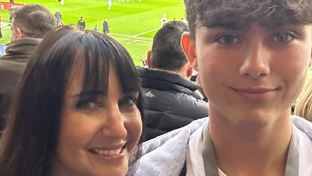 BBC presenter Lucy Owen on the left who is smiling next to her son Gabriel. He is wearing a white t-shirt and grey hoody. Behind them is the Etihad Stadium