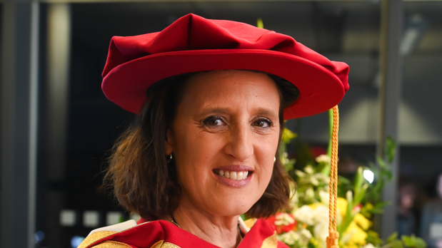 A headshot of Karen Pickering. She is wearing a red hat and a red gown having been given an honorary doctorate. She is smiling and looking into the camera.