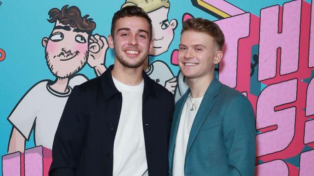 George Clarke and Max Balegde at the launch party for their podcast 'The Useless Hotline'. Both are stood on a red carpet, beside each other and against a in front of a backdrop that has podcast branding on.