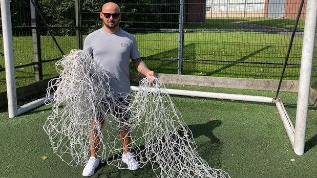 Rob Preece holding damaged nets. He is wearing a grey t-shirt, shorts, white trainers, and sunglasses. He is standing in front of a football goal