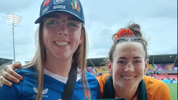 A woman with long mousy brown hair, glasses, a blue baseball cap and blue rugby shirt stands smiling next to a rugby player in a golden yellow top. The player has her brown hair scraped back and tied with an orange scrunchie. Behind  them is a rugby pitch, the opposite stand and floodlights