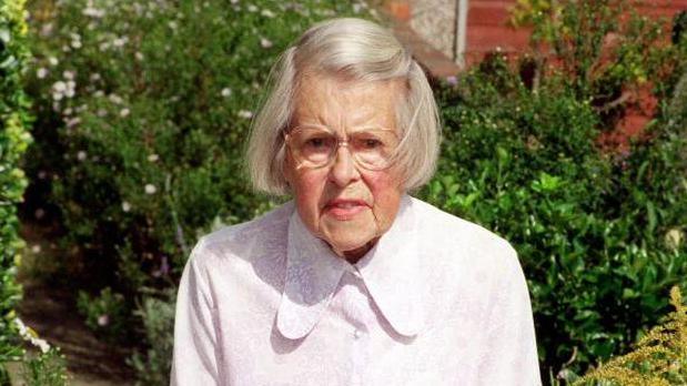 A colour photo of an elderly Melita Norwood. She has white hair cut in a short bob and is wearing a white shirt with a large 'Peter Pan' collar. She is standing in front of green shrubs and bushes.