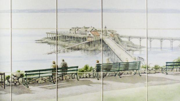 A close up image of the tiles showing an intricate painting of couples sat on benches overlooking Birnbeck pier and the old lifeboat station slipway into the sea. 