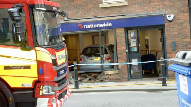 A longer-range view of the scene, which shows the car inside the branch of the bank. A red fire engine is visible in the foreground. 