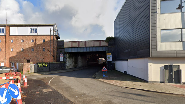 A road underneath a railway bridge. There are buildings on either side of the bridge and roadworks on the far left. 