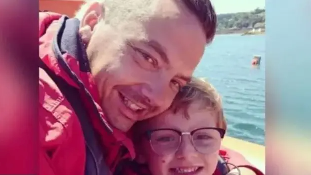 A man and son holding each other close and smiling to the camera in a selfie style, with the sea behind them. The young boy is wearing glasses and the father a large red coat.