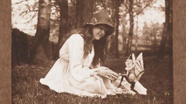 A black and white photo of a young girl with long hair sat on grass with trees in the background. She is wearing a white dress and a dark hat with a flower on the side and smiling whilst looking at a small mythical creature with wings and its foot in the air.