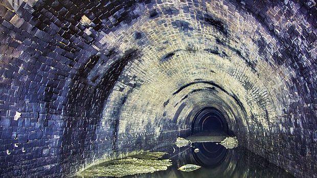 A disused railway tunnel wall with water on the floor reflecting the blue, black and white coloured stonework.