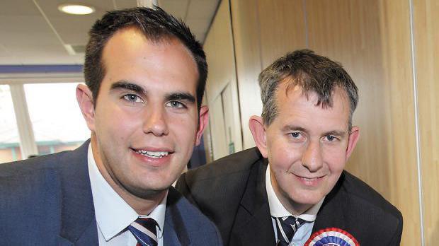 Luke Poots with short brown hair wearing a suit and a DUP voting pin standing beside Edwin Poots who has short brown and gray hair and wearing a dark suit. They are both smiling.