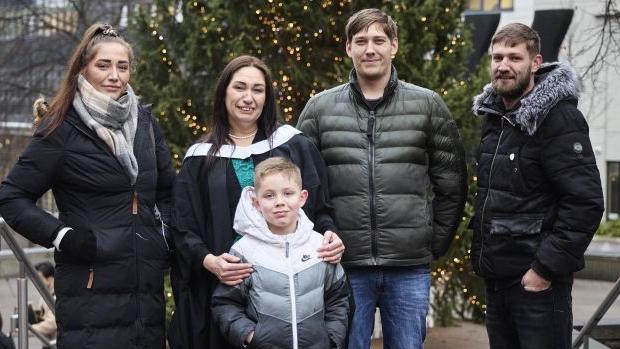 A young woman in a large puffer coat stands to the left of a woman in a graduation gown who has her arms on a young boy in a grey jacket. To the right of them stand two men, one in a green puffer coat, the other in a black jacket. Behind them is a Christmas tree.