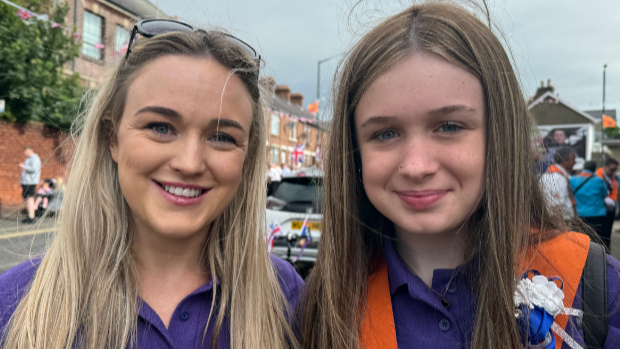 Cheryl Brownlee has long blonde hair, a purple top on and sunglasses on her head. She is smiling into the camera beside her daughter Brooke who has long brown hair and is wearing purple too with an orange sash. 