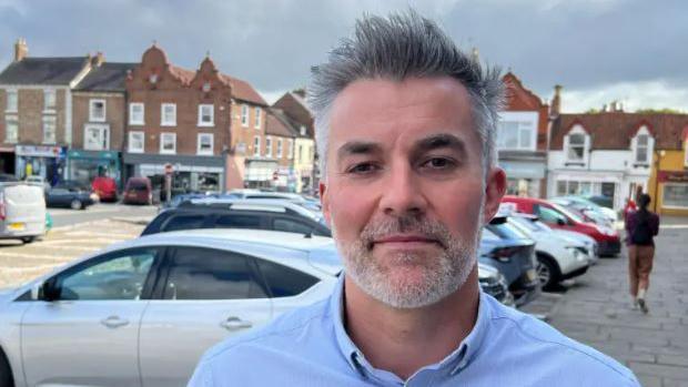 York and North Yorkshire Mayor David Skaith, stood in a car park in the county. He has a blue shirt and has short black and grey hair, with a short grey beard. Rows of cars are parked behind him. 