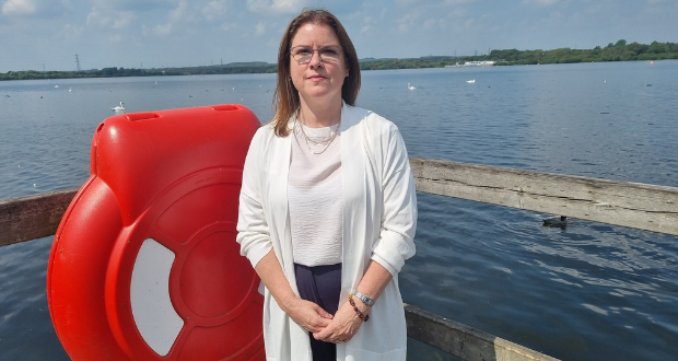 A woman with dark hair past her shoulders stands in front of a body of blue water. A fence bar can be seen behind her, grey, with the rest plastic of a life ring to her right. She wears transparent-rimmed glasses and a white top over a white top. On the water in the distance, swans can be seen.