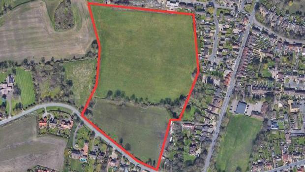 A bird's-eye view of the area that has been proposed for development. The space which is outlined in red currently is field space with several trees. 