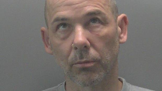 A custody image of Gavin Bainbridge - a man with a shaved head and a grey stubble beard. His eyes are looking up towards the ceiling.