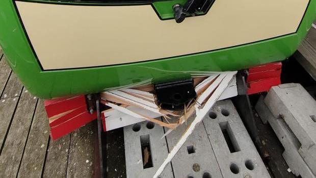 Shards of painted white wood from the train's front can be seen hanging from its body. The train is stationary on the tracks.