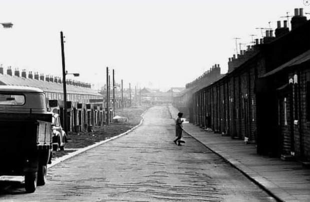 A black and white image of rows and rows of terraced miners' houses with a van and a single car parked up and a woman carrying a brush walking across the road.