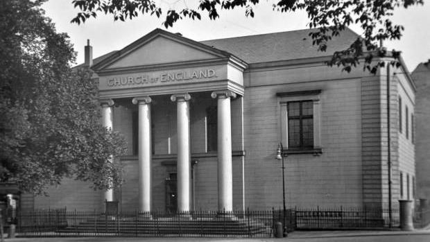 This photograph shows the English Church in Hamburg after its reconstruction in 1947. A large white/cream stoned church is pictured here in a black and white photograph. With four large columns, it's an imposing church surrounded by black railings. 