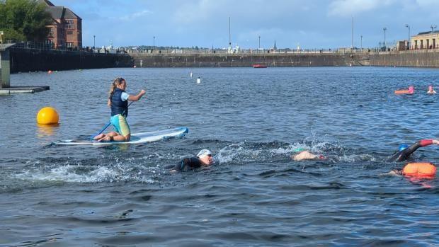 A man in a swimming hat can be seen swimming in the centre of the picture. A female warden is following nearby, kneeling on a paddleboard.