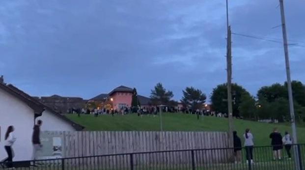 A number of young people can be seen congregating on a grassy embankment near a large pink building. Telephone wires and a picket fence can be seen at the bottom of the hill close to a white building.